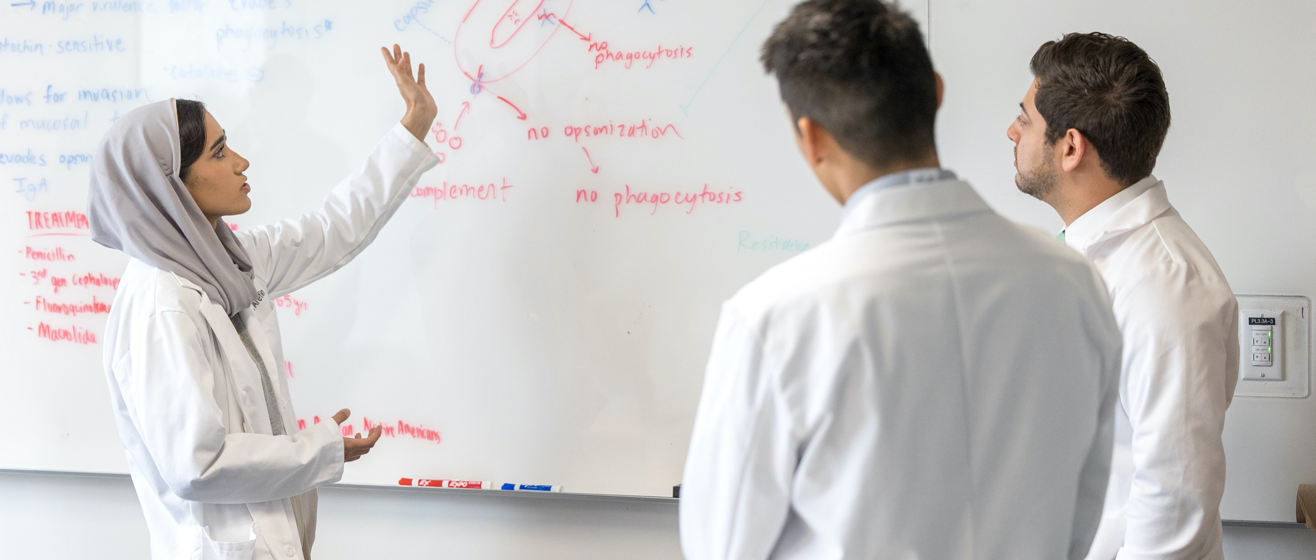 A female Dell Med student discusses a medical diagram drawn on a whiteboard. Two other Dell Med students, both male, look on.