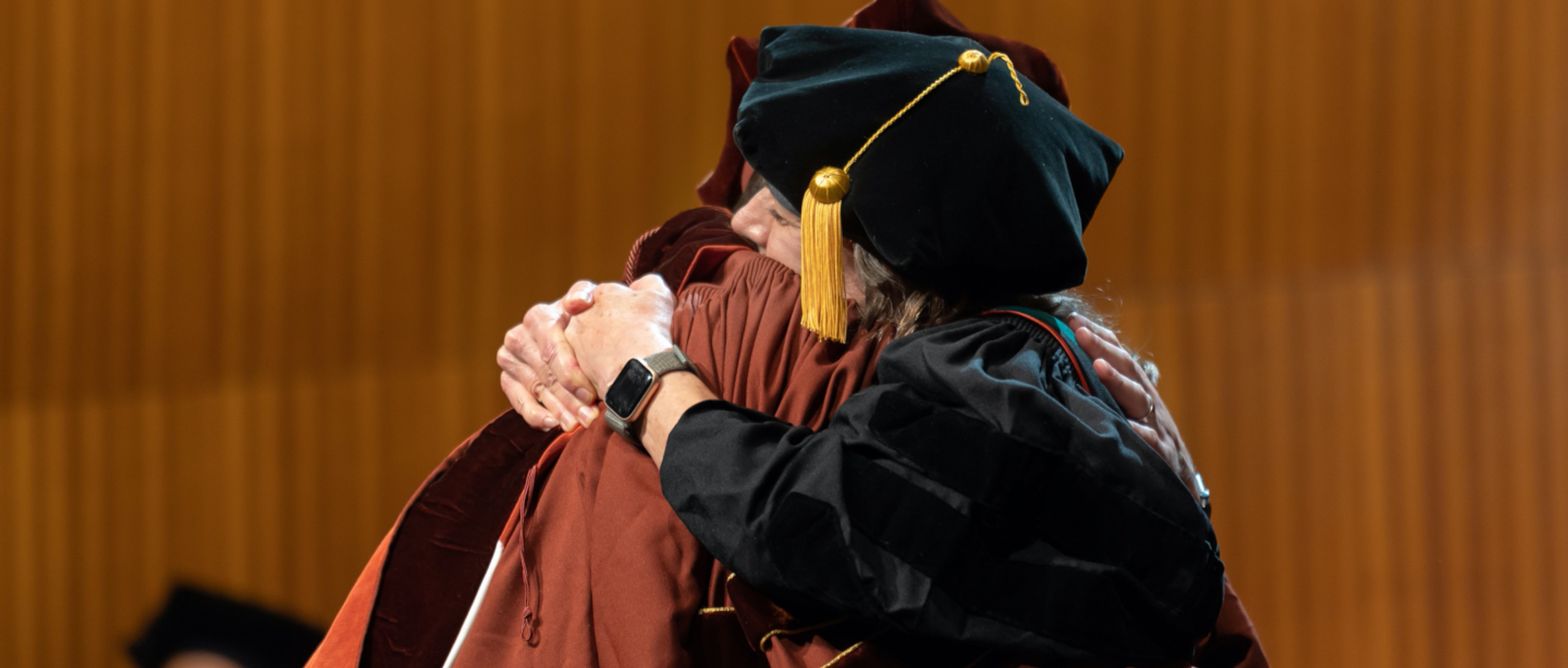 A Dell Medical School faculty member hugs a graduating student. Both faculty member and student wear respective graduation ceremony regalia.