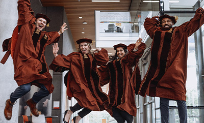 Four students from Dell Med's Class of 2024 — each wearing graduation regalia — jump for a celebratory portrait inside the Dell Med campus.