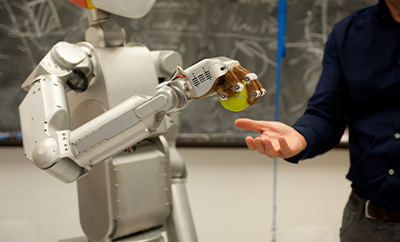 A robot holds a tennis ball over a person's hand that anticipates receiving it.