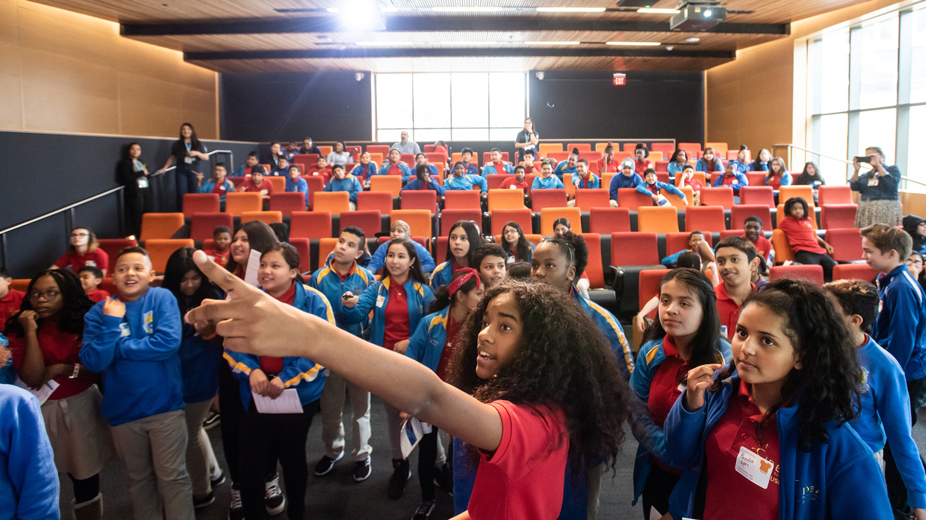 A female middle school student pointing at a projector screen while the rest of her class stands and sits behind her in an auditorium.