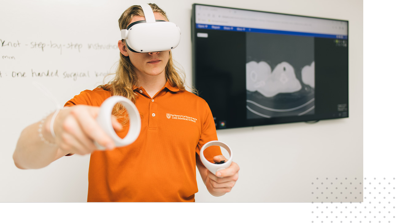 A Dell Med student wears a virtual reality headset with accompanying handheld devices. He stands in front of a whiteboard and monitor.