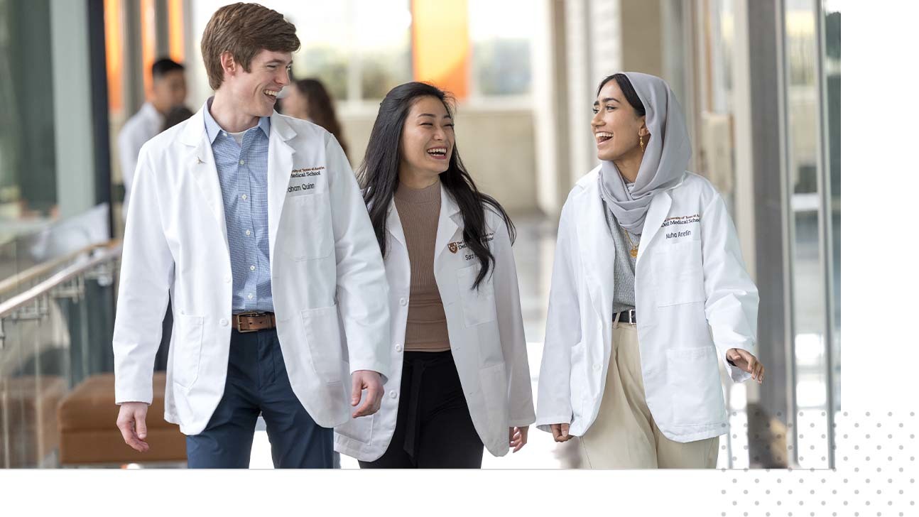 Three medical students, each wearing white coats, walk together inside the Dell Medical School campus smiling.
