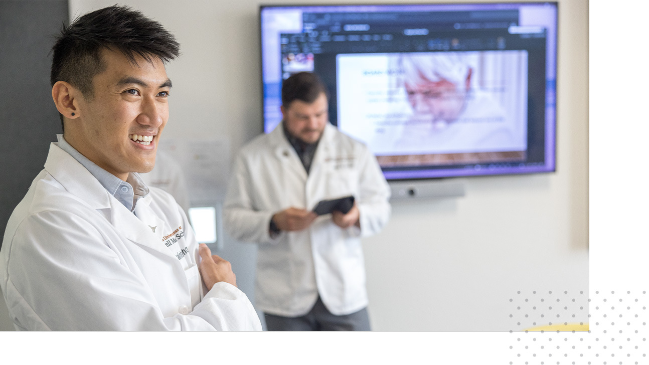 Two Dell Med students, both wearing medical white coats, stand during a small-classroom activity.