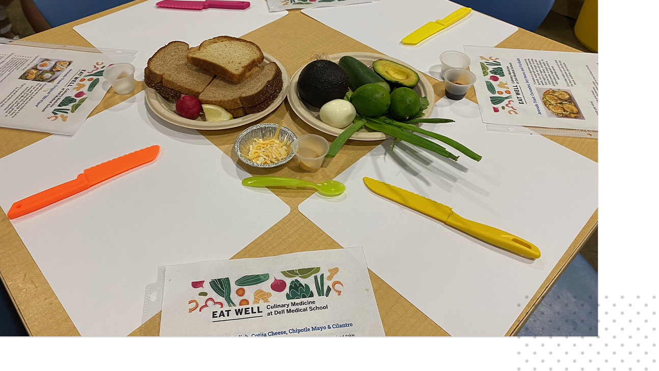 A table with four placemats and seats, and at each seat there is a recipe sheet. In the middle of the table are ingredients to make the recipe on the sheet.