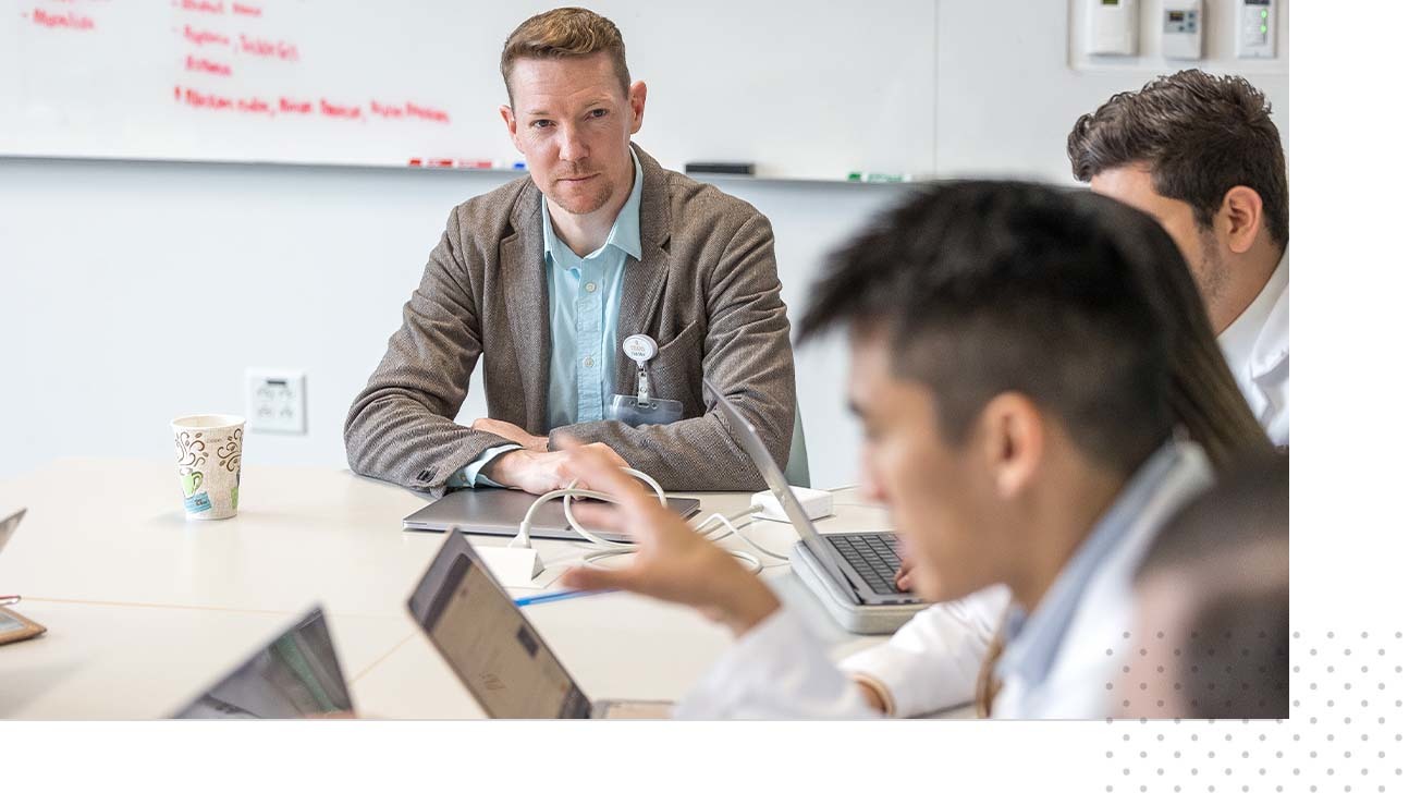 All sitting at a classroom table, a Dell Med instructor looks on at students working on their laptops.