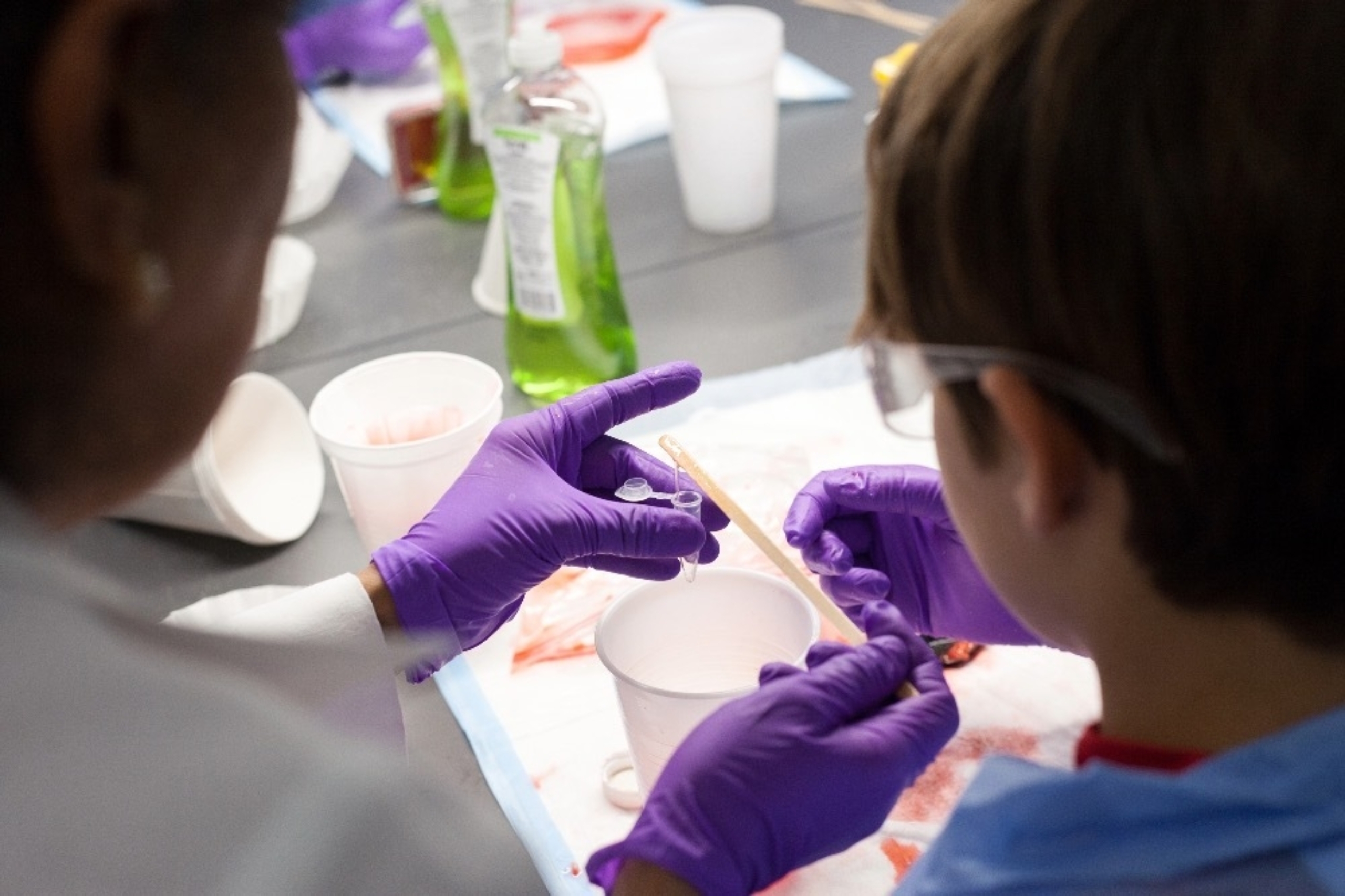 Two individuals wearing rubber gloves conduct as science activity with liquid and a styrofoam cup during Explore UT.