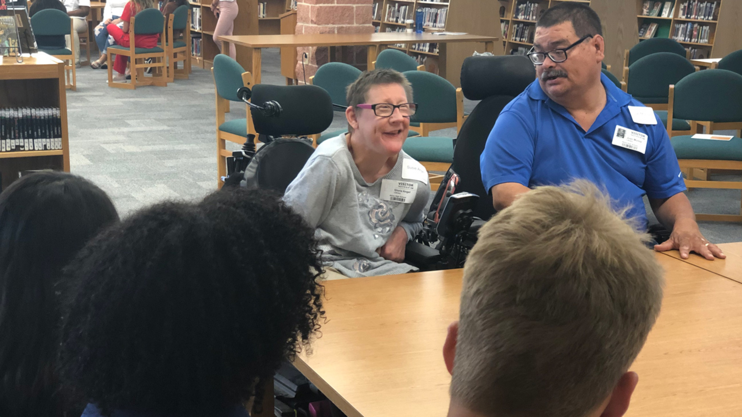 A man and a woman in wheelchairs speak to a group of students sitting at a table.