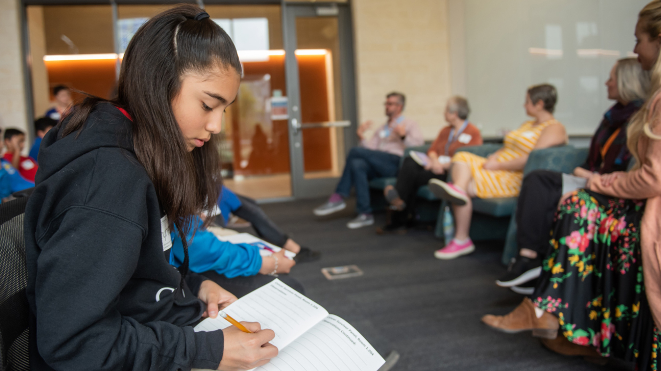 A middle school girl sitting in a chair taking notes during a panel discussion.