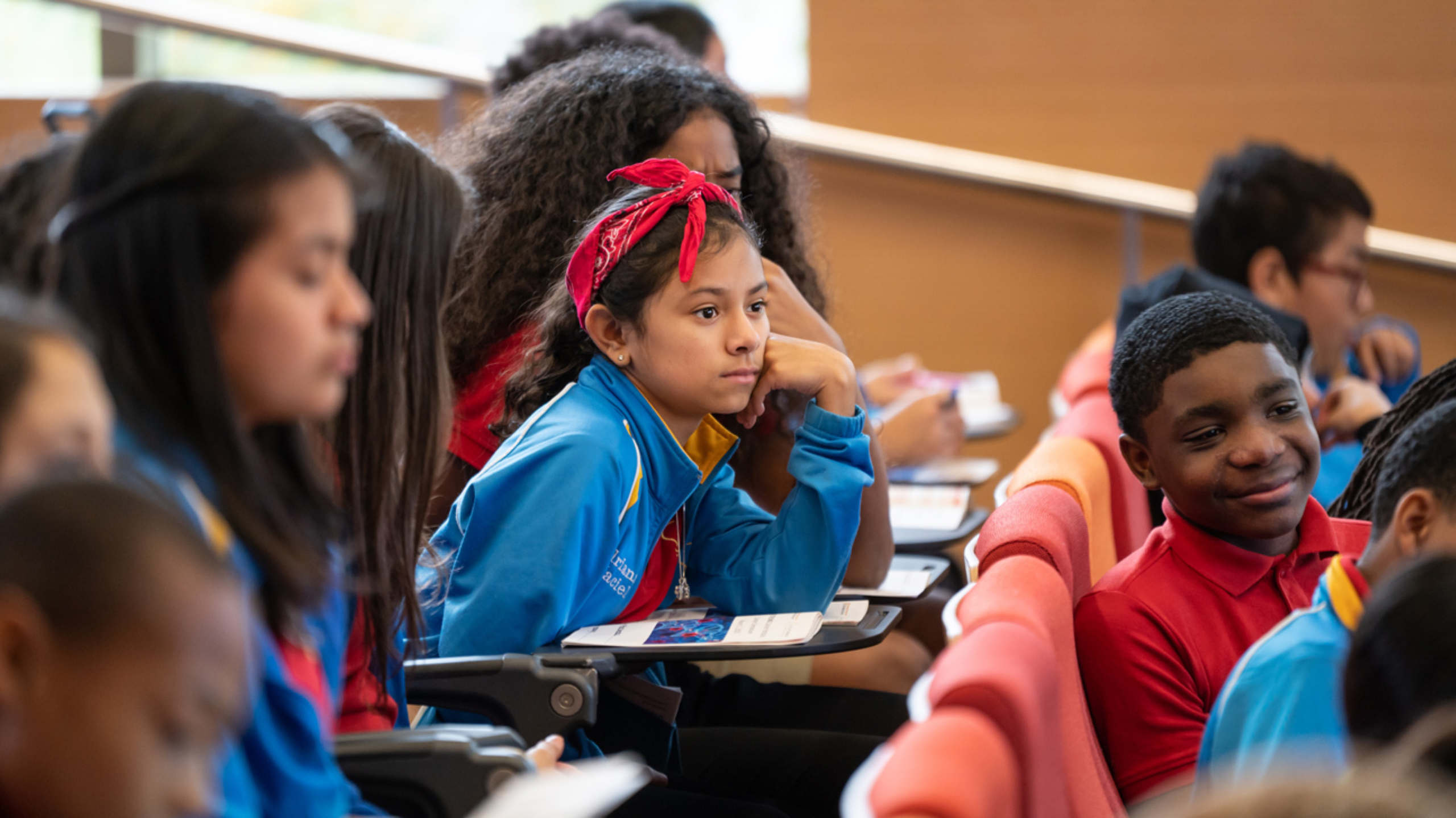 A middle school girl sitting in an auditorium with her class paying attention to a lecture with her head resting on her hand.